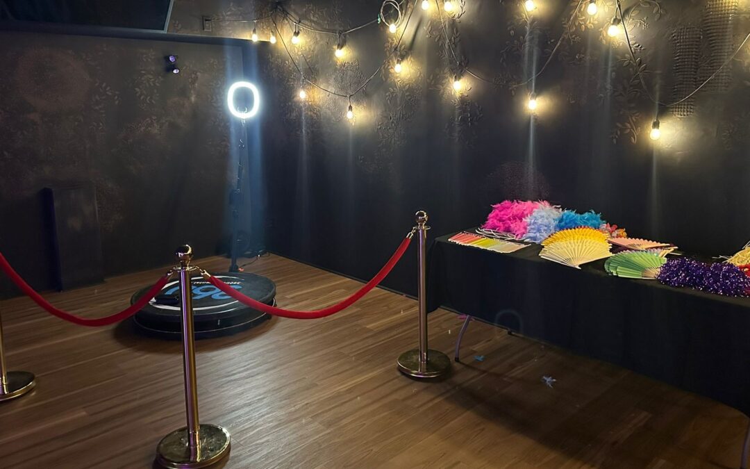 A 360 video booth platform on a wooden floor in a dark room, with red rope stanchions set in front of it, a string of warm bulb lights running across the patterned wall behind and a props table of feather boas and paper fans alongside