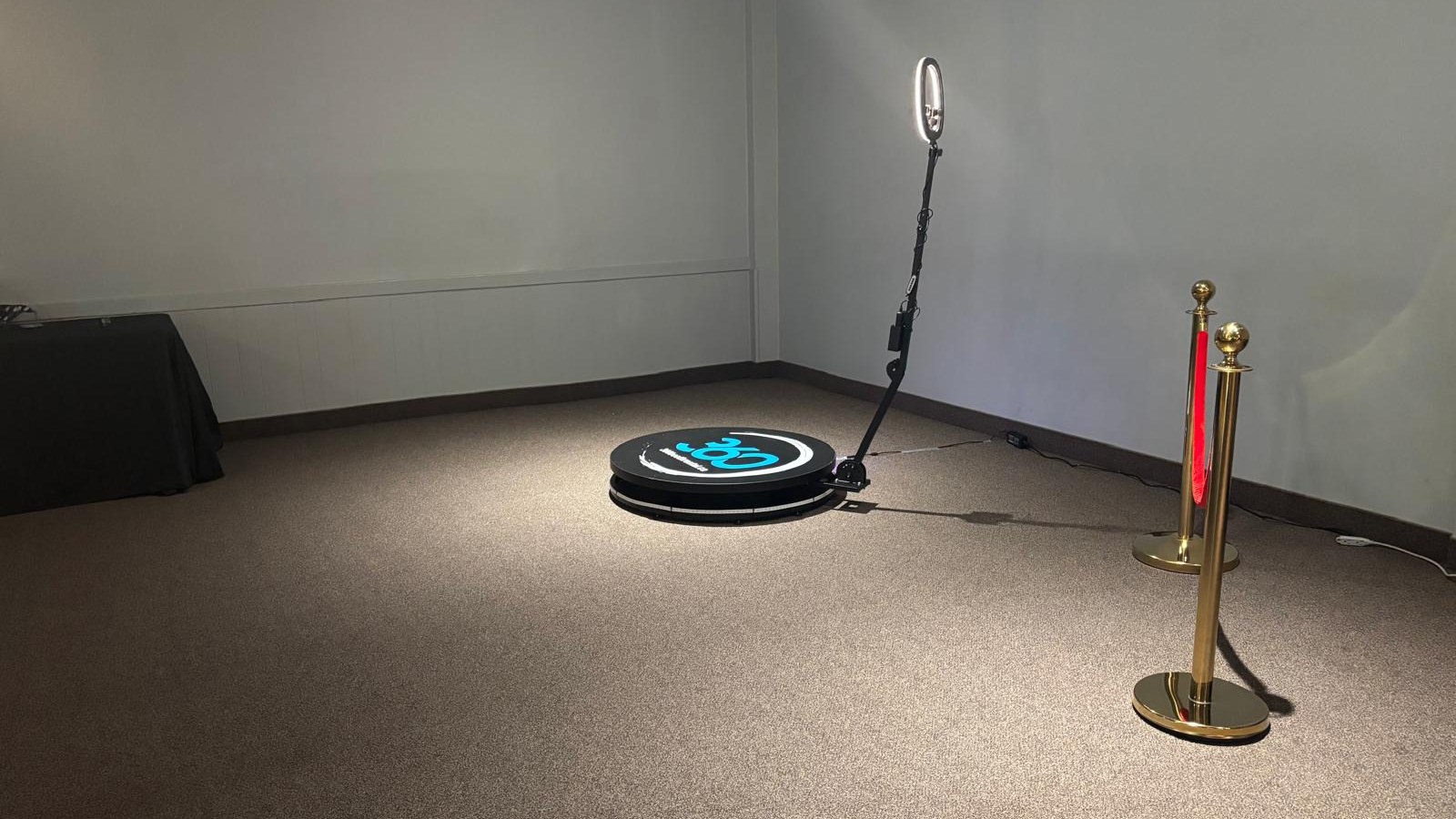 A 360 video booth platform and its ring light standing on their own on the carpet of a plain pale room, with a rope stanchion to one side and clear floor on every side of the platform