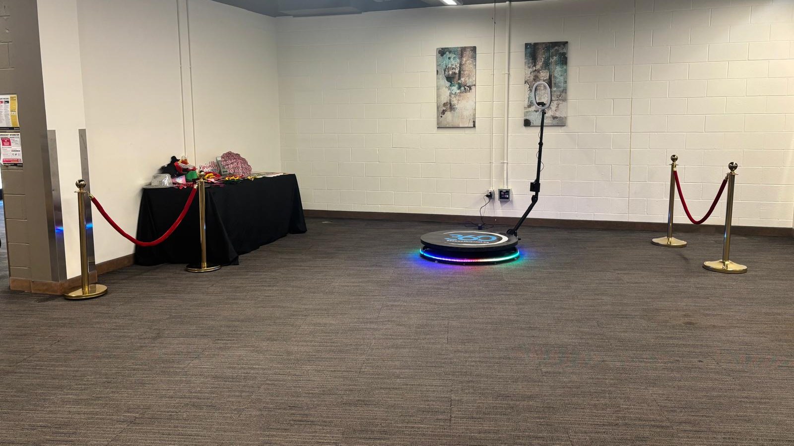 A 360 video booth platform with its light ring lit, four rope stanchions standing around it and open carpeted floor running away in front, with a draped props table against the wall beyond