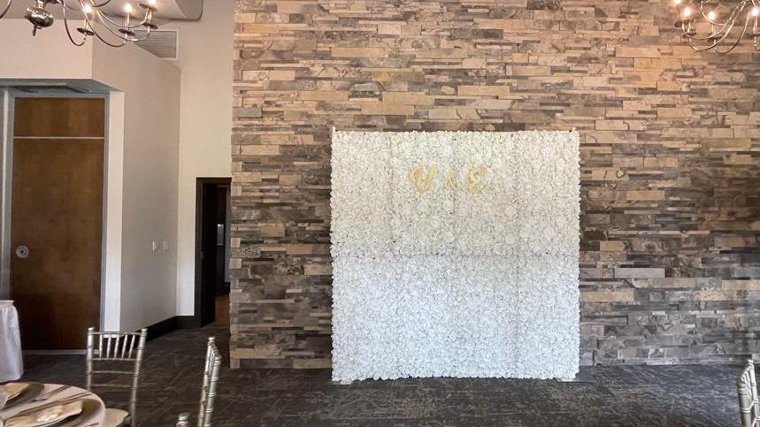 A white flower wall standing against a stacked stone feature wall in a banquet room, with patterned carpet, two chandeliers, exposed ducting and set tables around it
