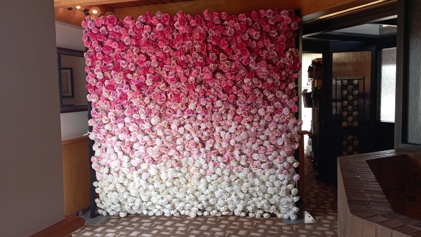 A pink and cream ombre rose flower wall standing in a room with a timber plank ceiling and beam, on patterned carpet beside a brick hearth