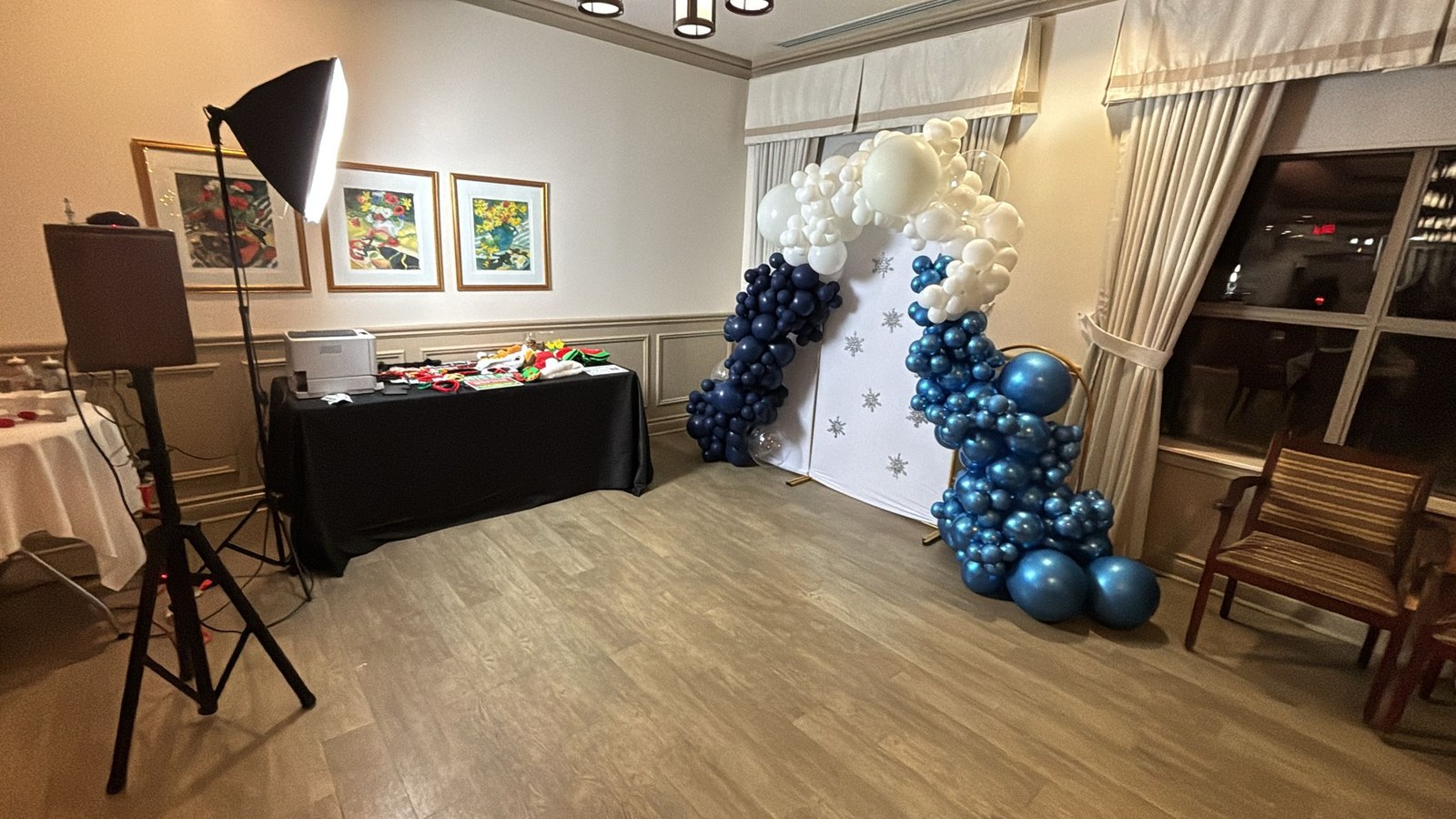 A navy, chrome blue and cream balloon garland framing a white backdrop printed with silver snowflakes, with party props laid out on a black skirted table and a softbox beside it in a panelled function room