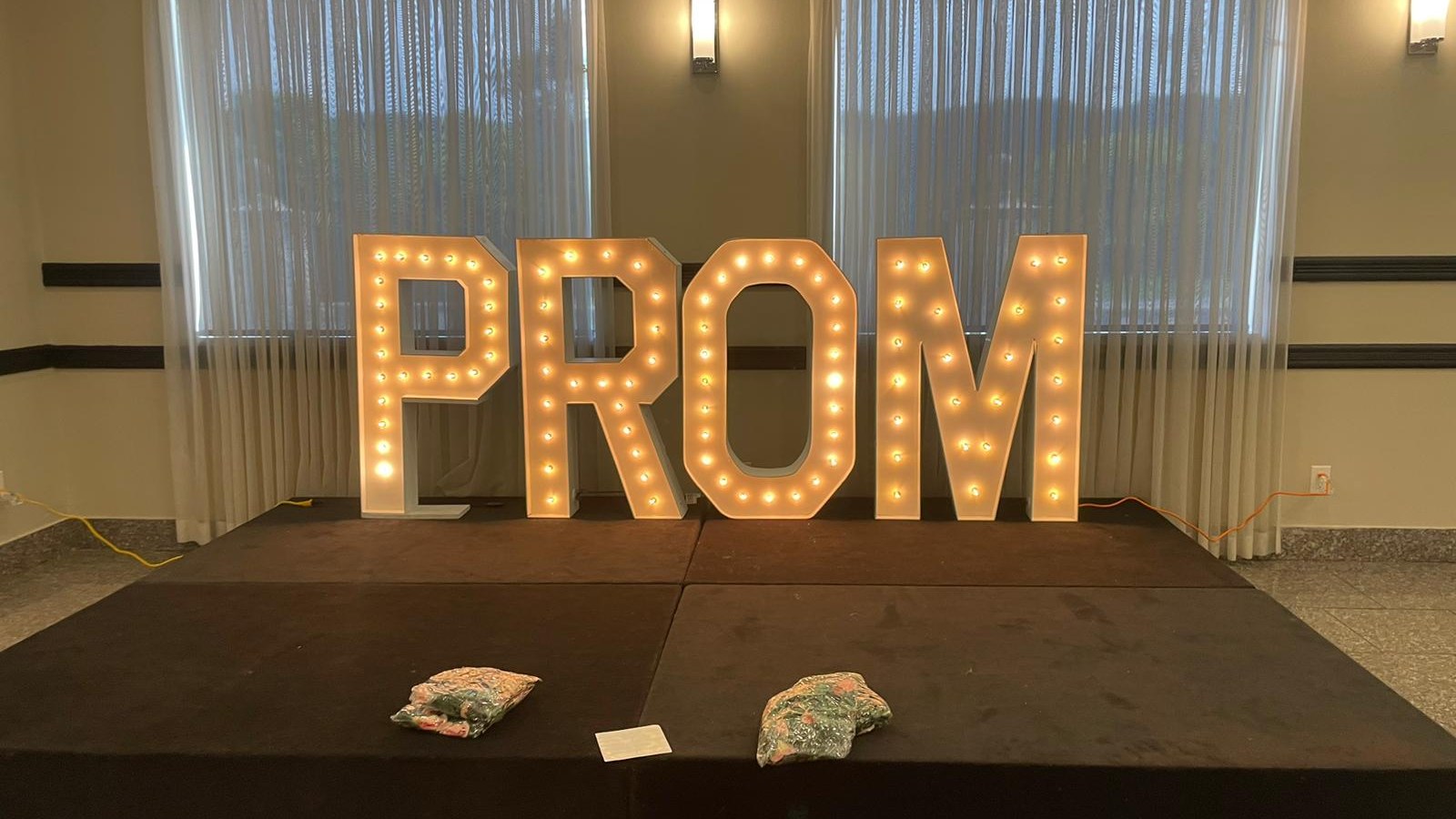 Lit marquee letters spelling PROM standing on a dark skirted stage with two bagged bundles, a card and extension cords on the boards in front of them