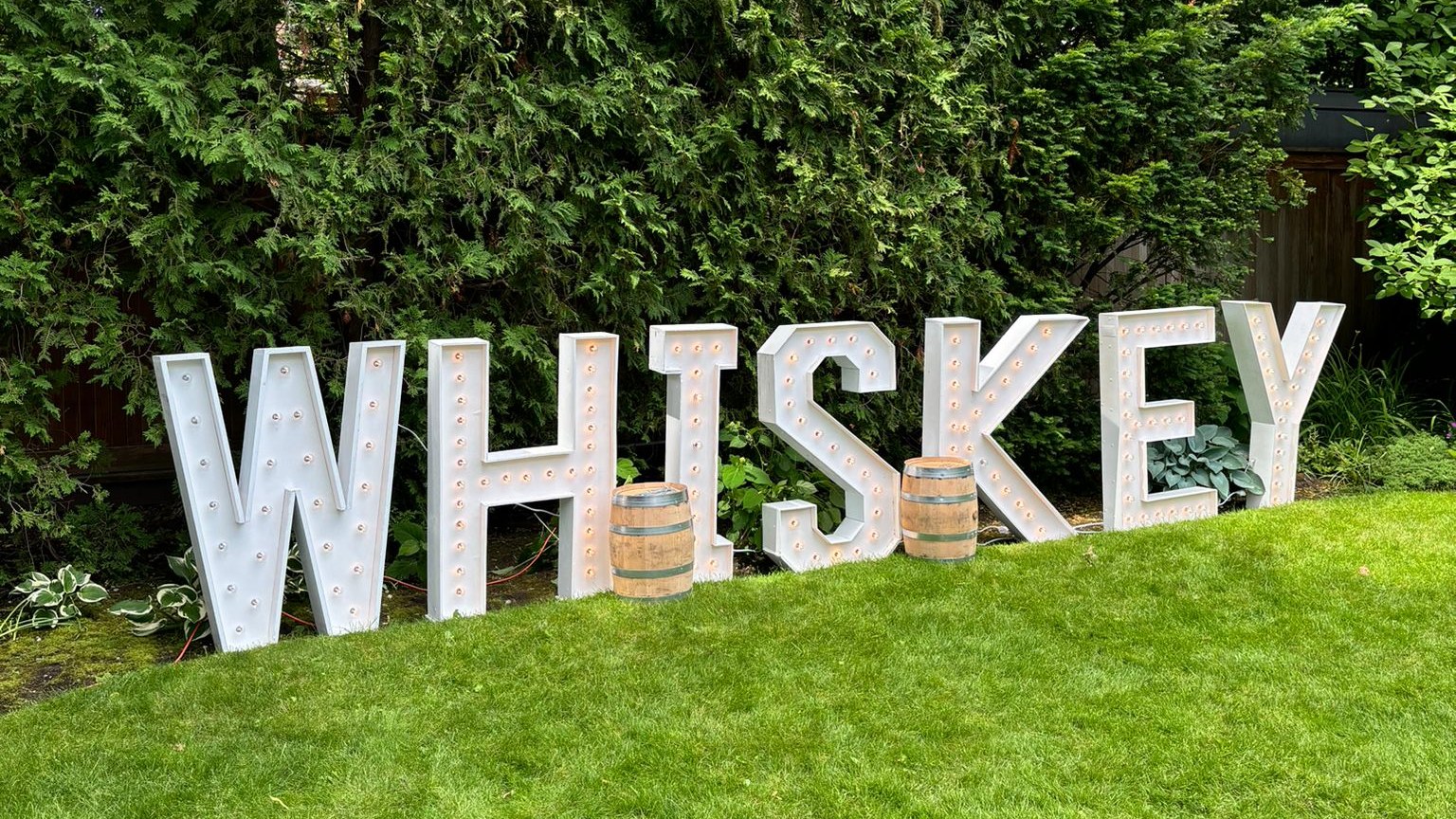 Lit marquee letters spelling WHISKEY standing on a lawn in daylight in front of a tall cedar hedge, with two wooden barrels between them and a cable running along the grass