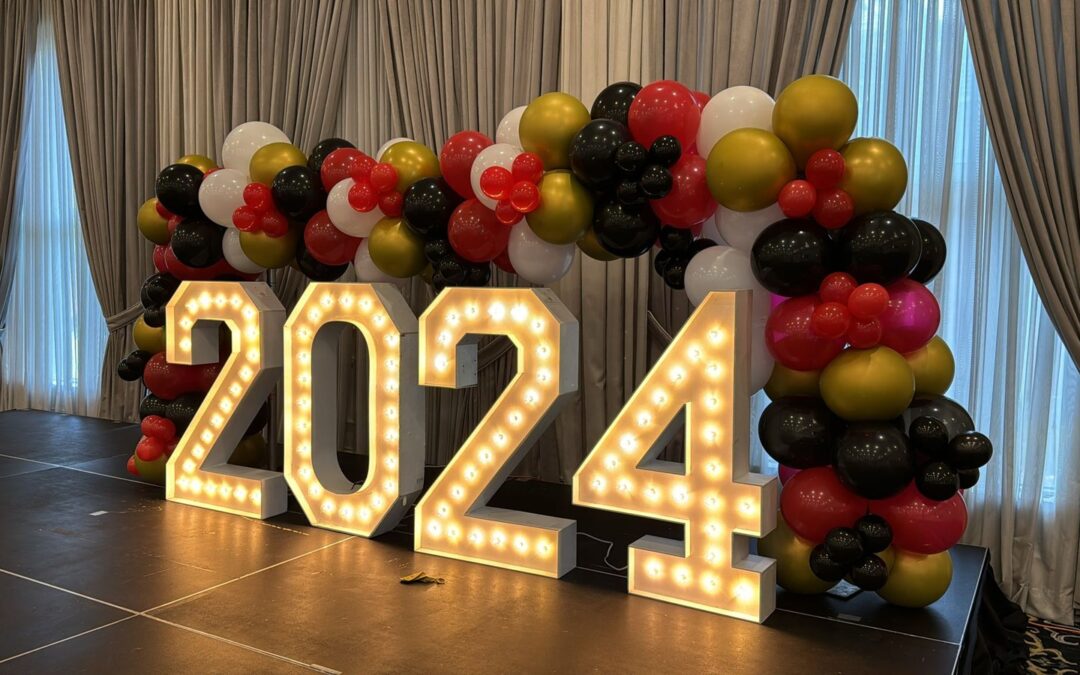 Lit marquee numbers reading 2024 standing on a low stage under a red, black, gold and white balloon garland against full length curtains