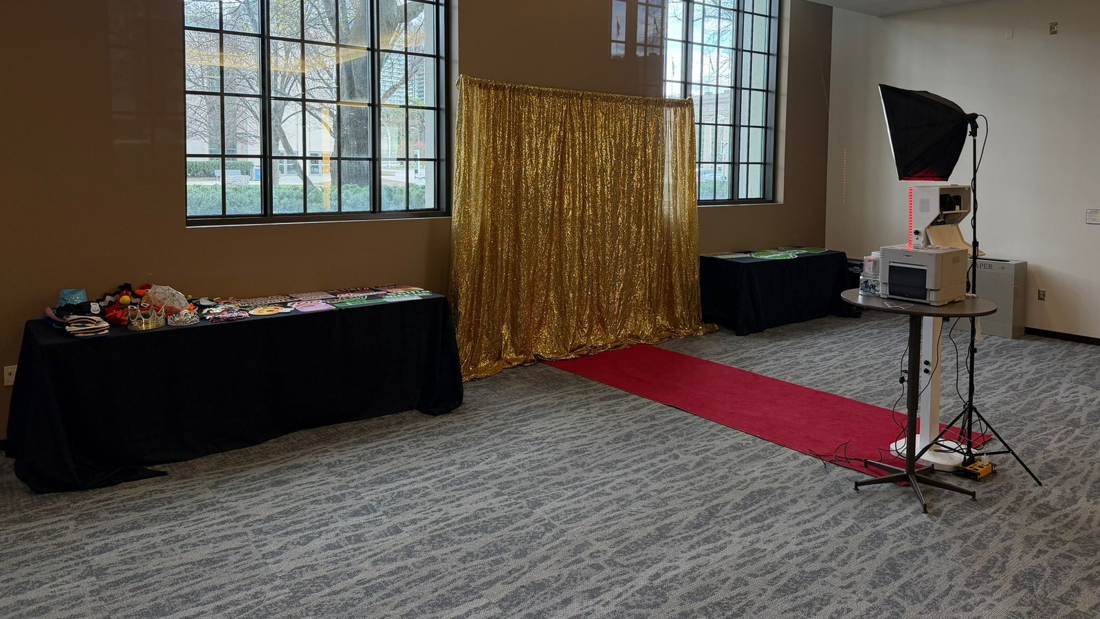 A gold sequin backdrop and a red carpet runner set up in daylight under tall windows, with a long props table on one side and a photo booth, softbox and printer on the other