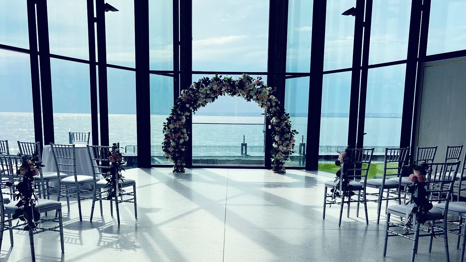 A floral moon gate archway standing against a tall glass wall, with rows of chiavari chairs in front of it and open water on the horizon behind