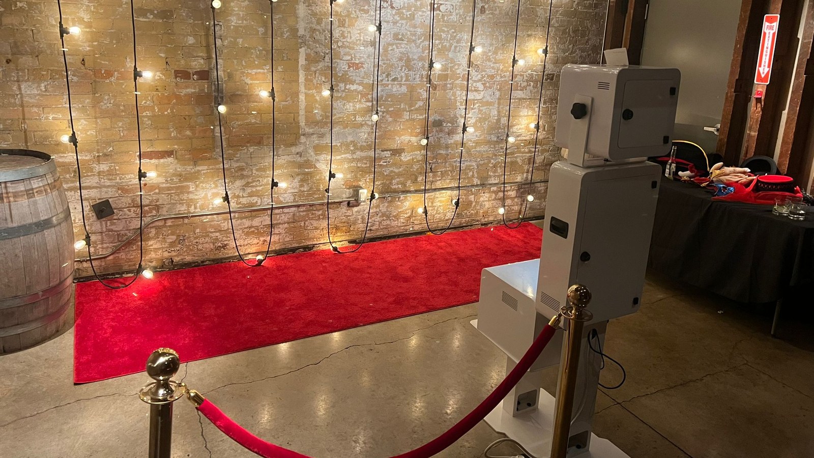 A red carpet runner laid indoors along an exposed brick wall hung with bulb lights, with a gold stanchion and red rope across the front and a photo booth beside it
