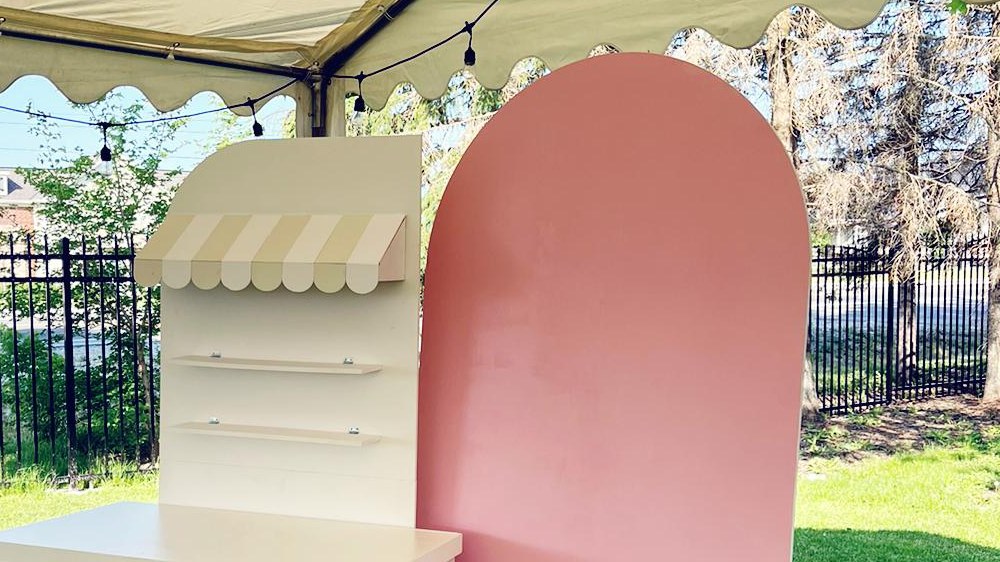 The underside of a cream marquee canopy over a lawn, with festoon lights strung beneath its scalloped edge and a cream candy cart standing at the edge beside a pink arch panel