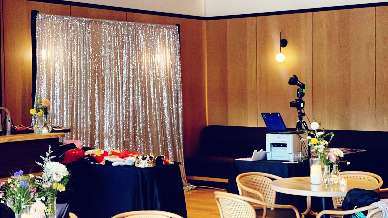 A champagne sequin backdrop lit warm in a dark panelled room, with a props table, the booth camera on its stand and a photo printer set up at the side