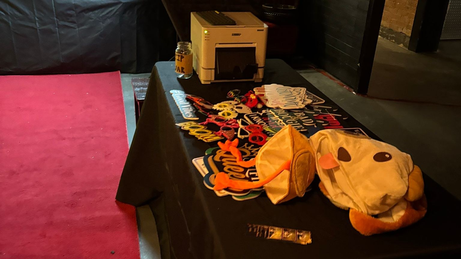 A props table holding a photo printer, prop signs and a plush hat beside a red carpet runner and the dark wall of a photo booth, in a low lit brick walled bar