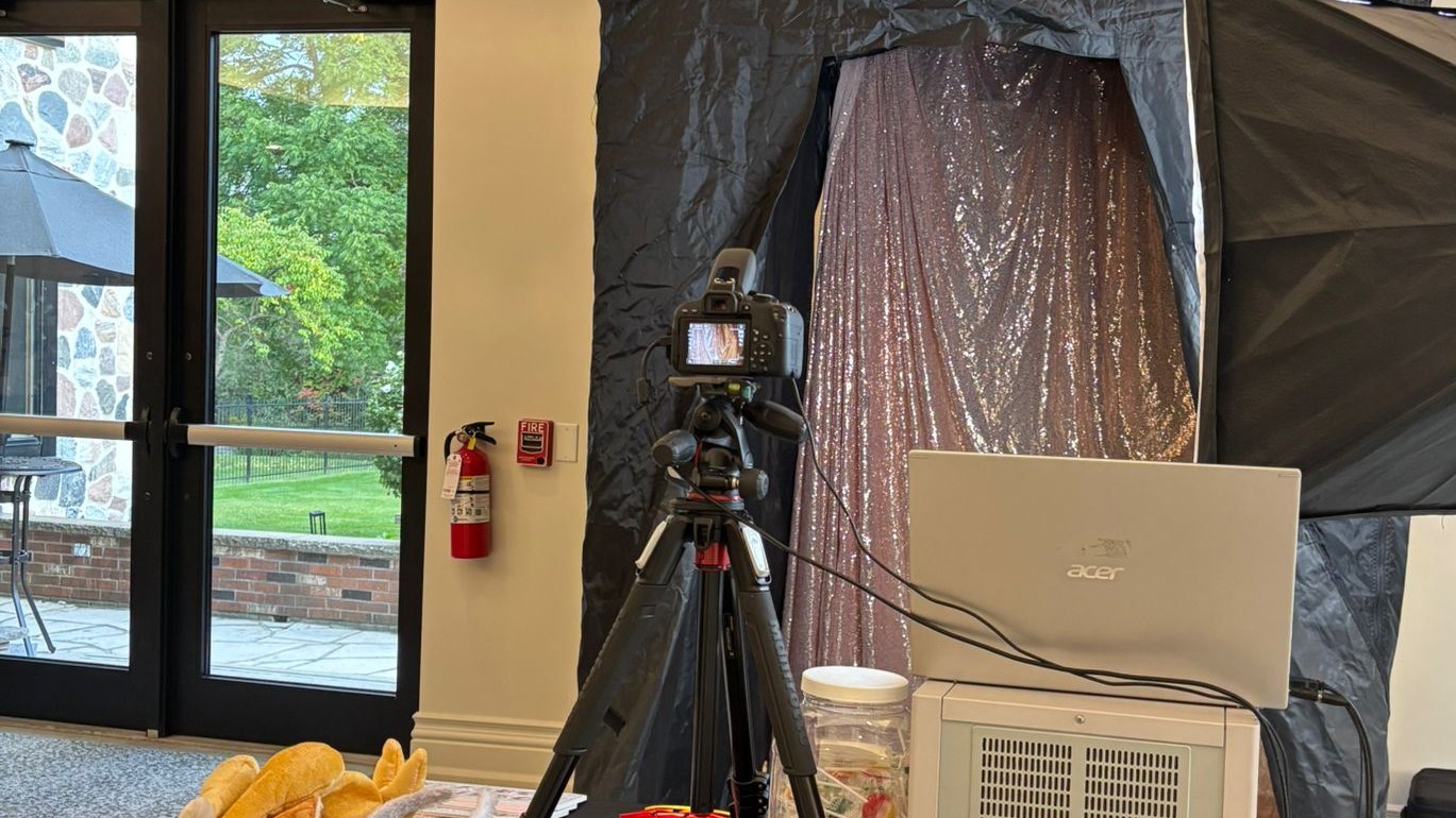 An enclosed photo booth shell with a rose gold sequin backdrop hung inside it, the camera on a tripod and a laptop and printer alongside, set up in a plain room with glass doors onto a terrace