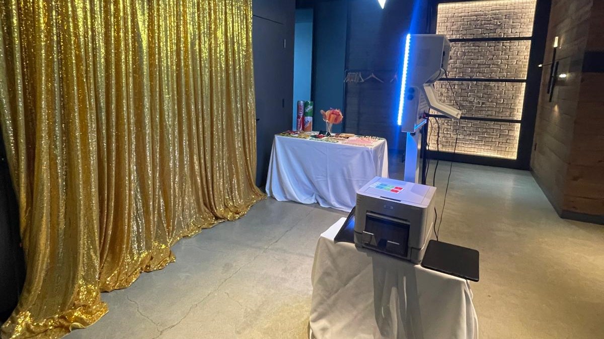 A photo booth pod with a lit blue column and its printer on a draped table facing a gold sequin backdrop in a dark walled room, with a props table of giant foil drinks cans and a lit brick panel beyond