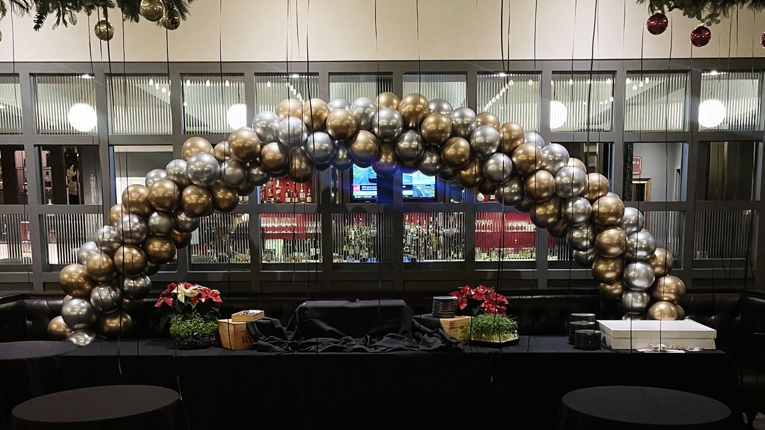 A chrome gold and silver balloon arch built over a long black skirted counter in front of a glazed back bar, with poinsettias, a Christmas garland above and black cocktail tables on an empty floor