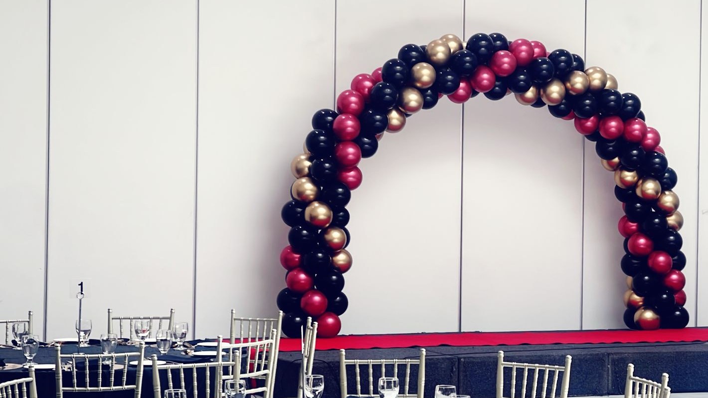 A black, red and gold balloon arch standing on a stage against a white panelled wall, with both legs based on a red carpet strip and gold chiavari chairs at tables in the foreground