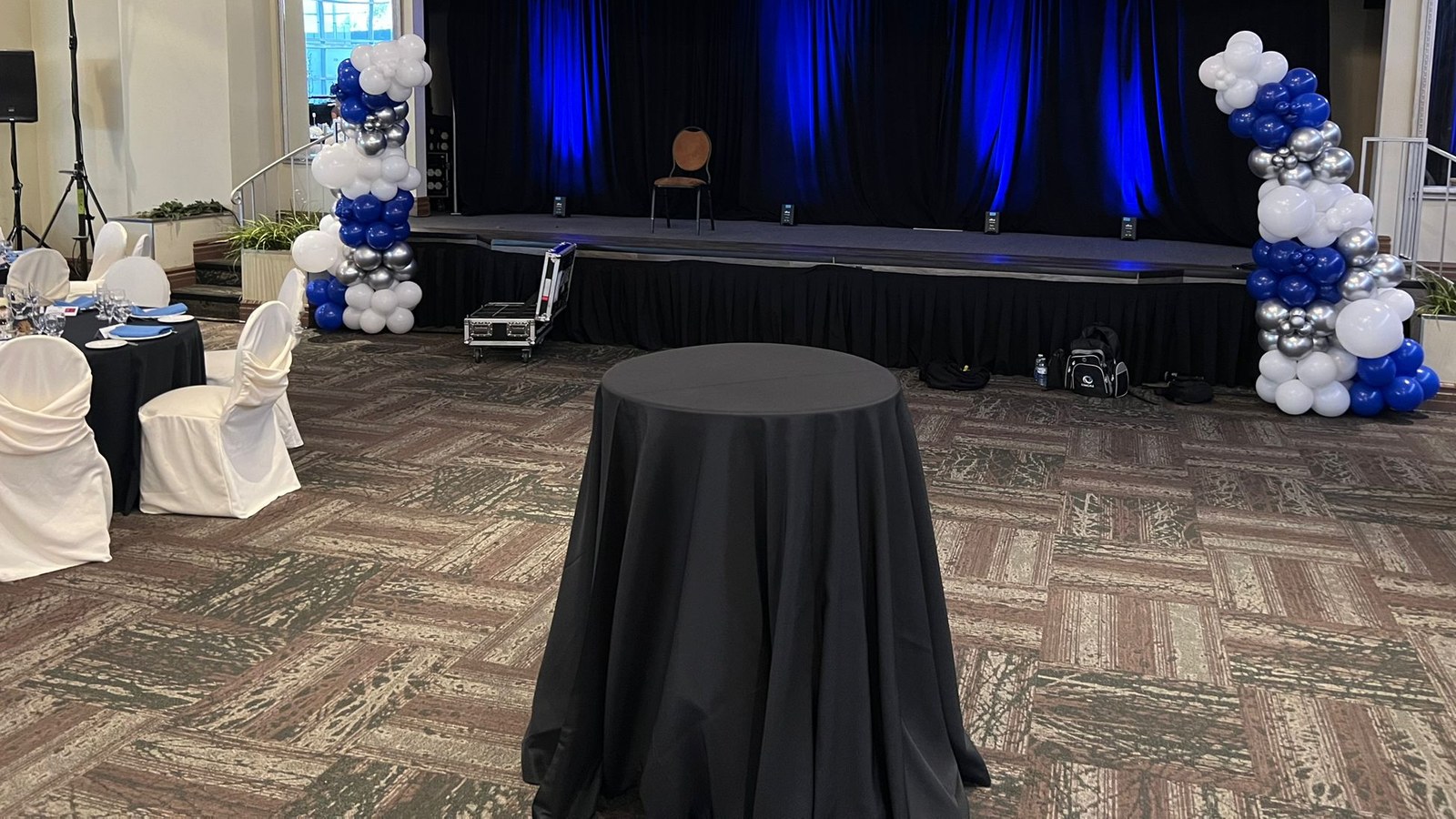 Two blue, white and silver balloon columns either side of a low stage lit blue, with a black skirted cocktail table on the floor, covered chairs along one wall and a road case and bag still standing by the stage