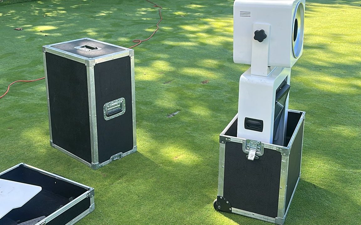 Two black flight cases standing on mown grass with a photo booth pod lifted half out of one of them and a third case open flat at the edge of the frame