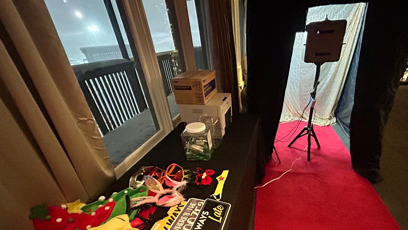 A red carpet runner leading into an enclosed photo booth with a pale sequin backdrop and a screen on a tripod, set at the edge of a dark room whose windows look onto snow and a lit deck rail, with a props table in the foreground