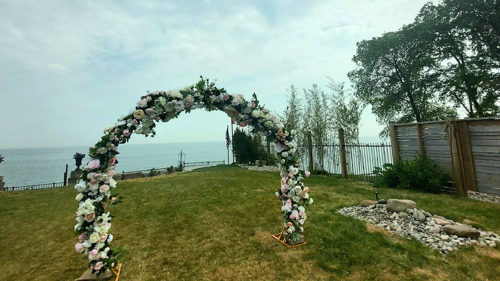 A floral moon gate arch standing alone on a mown lawn under open sky with nothing above it, its gold feet weighted down with stone slabs