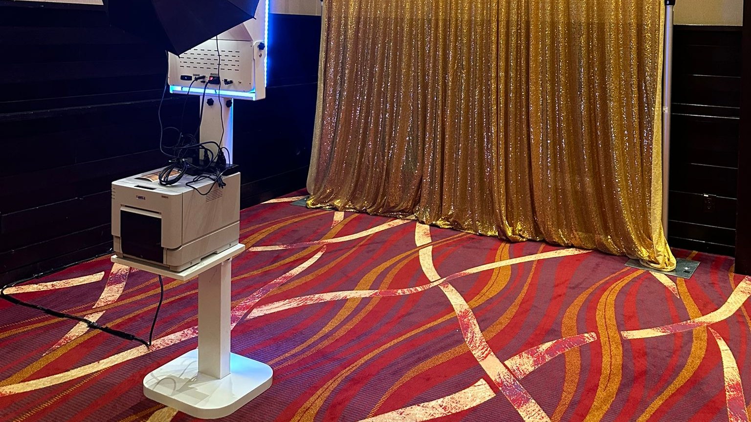 A photo booth pod with a lit blue column and its printer standing on a patterned red and gold banquet carpet beside a gold sequin backdrop whose hem rests on the floor