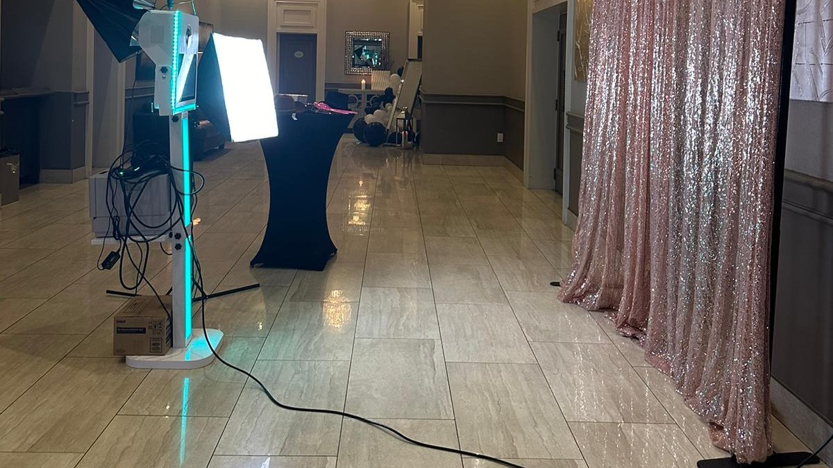 A photo booth pod with a lit turquoise column facing a rose gold sequin backdrop with its power cable run across a tiled function room floor