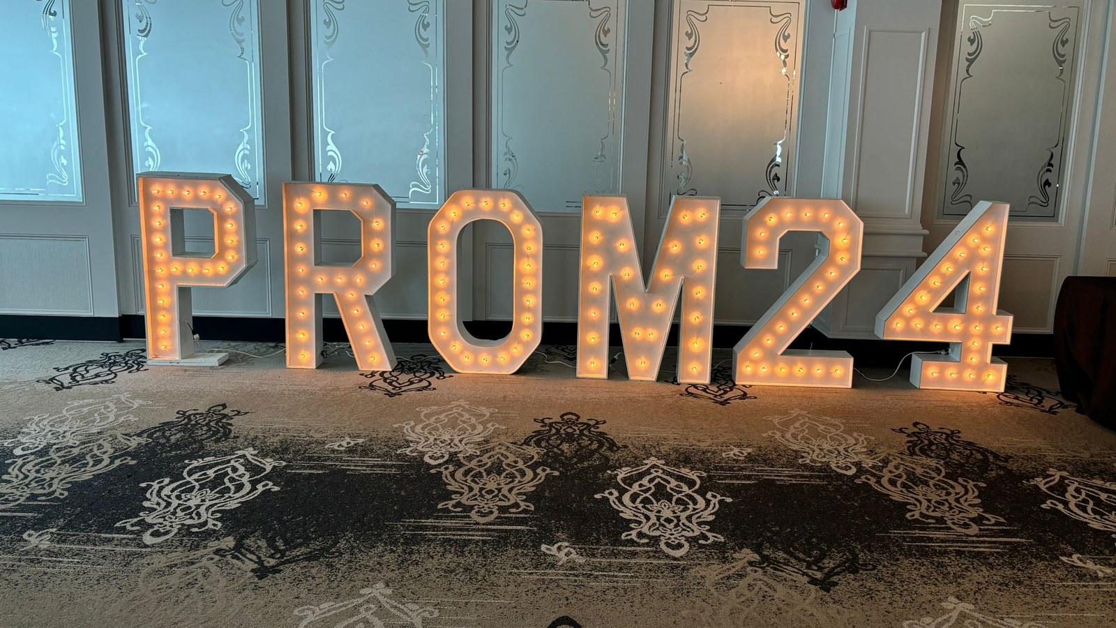 Lit marquee letters and numbers reading PROM24 standing on a patterned carpet along the etched glass and panelled wall of a wide room