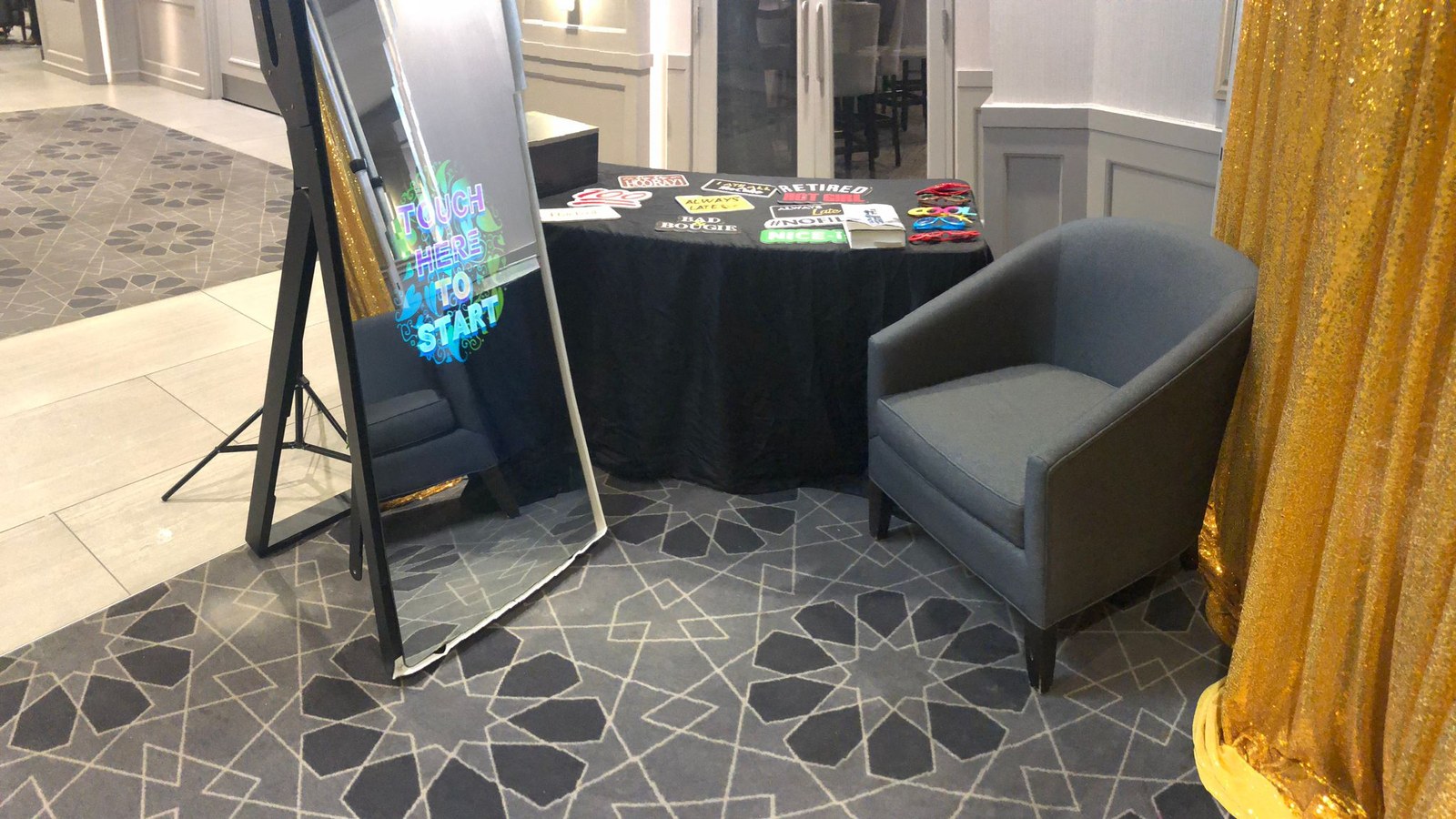A mirror photo booth set at the side of a carpeted function room beside a cloth covered props table, an armchair and the edge of a gold sequin backdrop