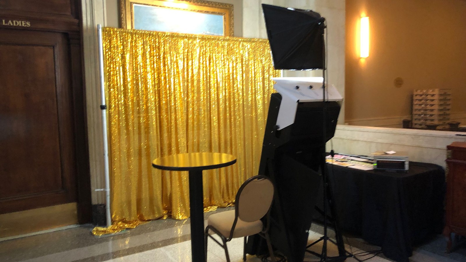 A mirror photo booth and its softbox light standing ready against a gold sequin backdrop in a panelled lobby, with a bar height table, a banquet chair and a draped side table holding the printer