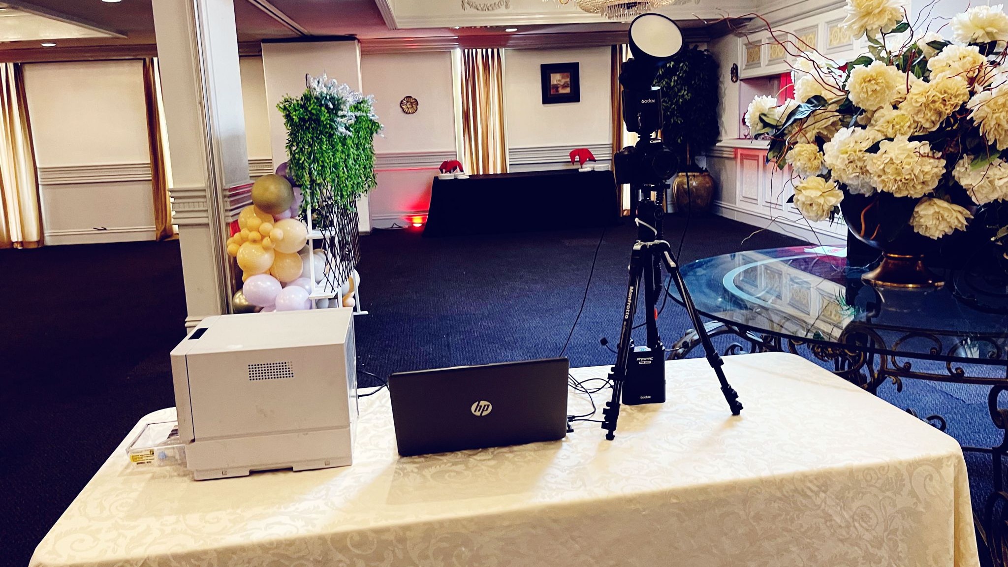 A photo booth printer, a laptop and a camera on its stand set up on a cloth covered table at the side of a function room, with a balloon column and a tall cream flower arrangement beside the table and a long draped head table across the room behind