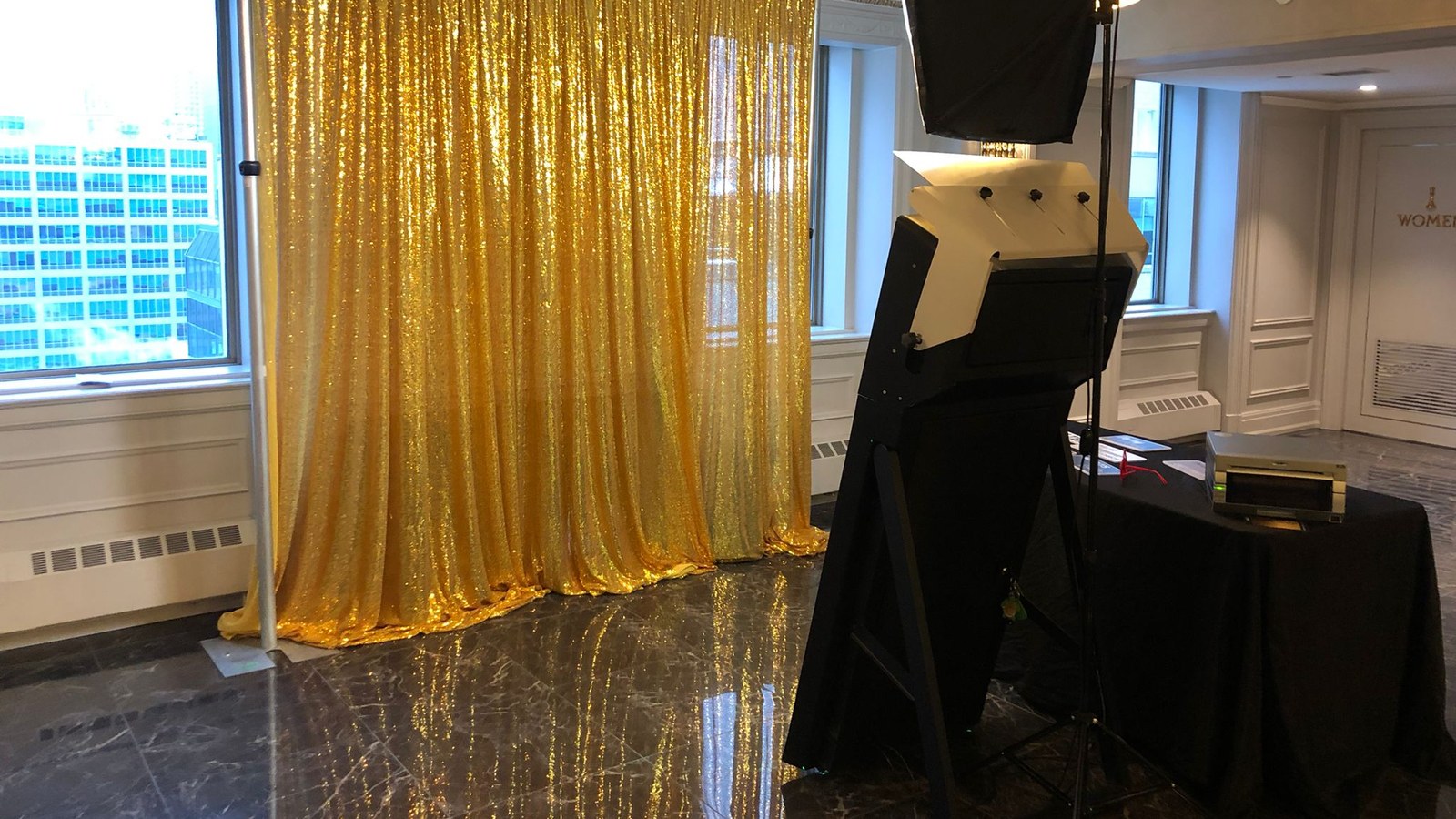 A mirror photo booth on its stand with a softbox light overhead, standing on polished marble beside a full length gold sequin backdrop, with a photo printer on a black draped table at the side